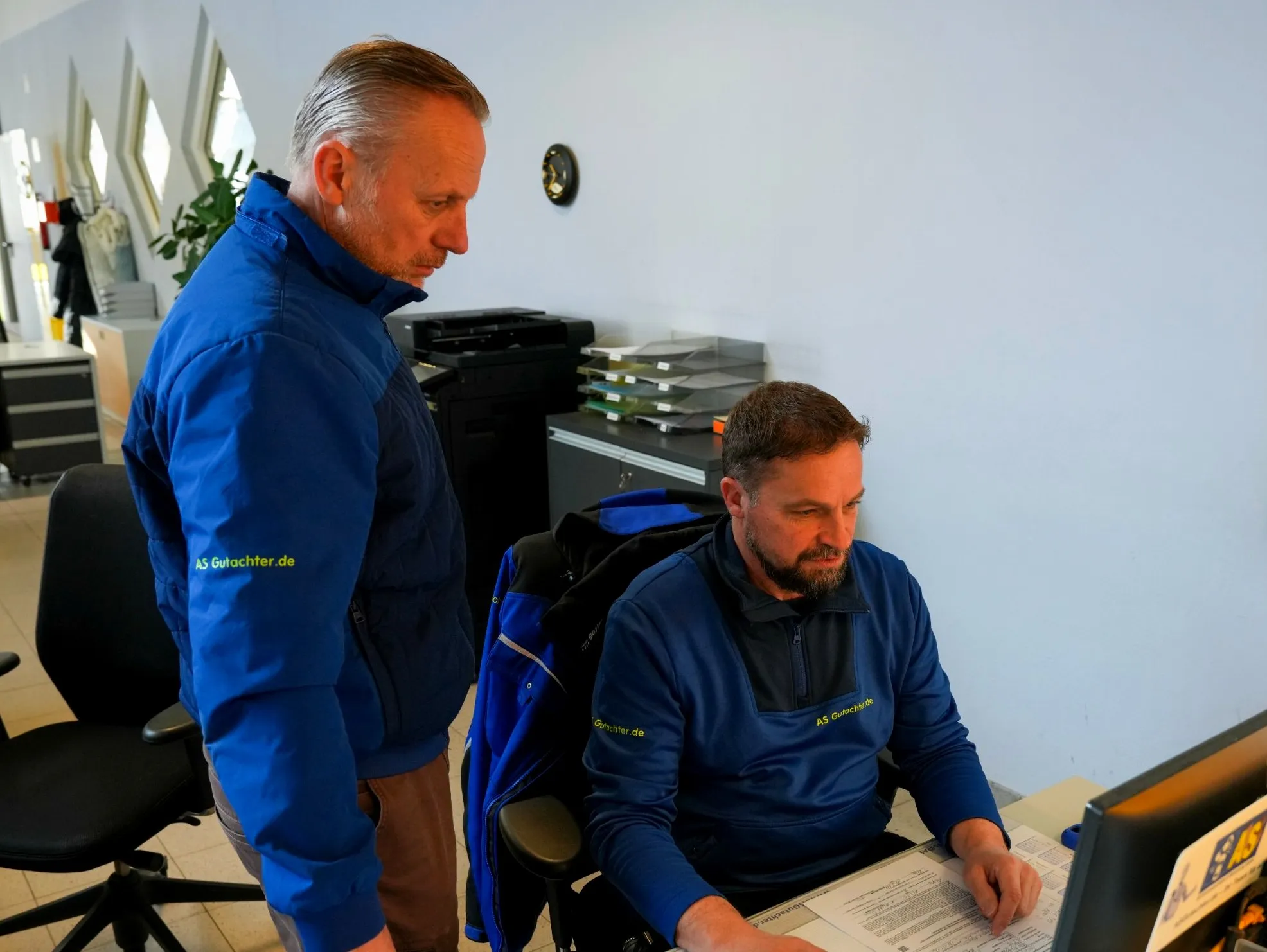 Alexander Ströher steht im Büro hinter Bernhard Rührmair, der am Schreibtisch ein Gutachten bearbeitet.